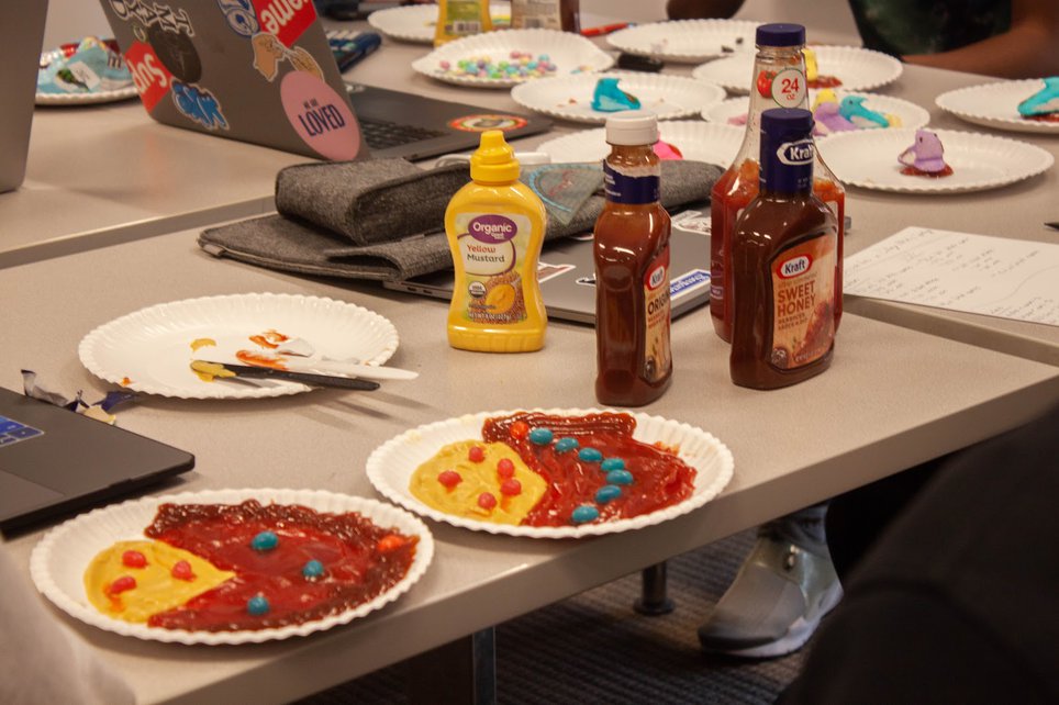 Plates of multicolored condiments rest on a table with bottles of mustard and ketchup.